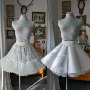 May include: Two white tulle skirts on dress forms. The skirt on the left is a layered tulle skirt with a white satin waistband. The skirt on the right is a single layer tulle skirt with a white satin waistband.
