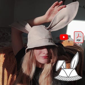 May include: A beige bucket hat with long bunny ears. The hat has a simple design and is made of a soft fabric. The hat is being worn by a person with long blonde hair. The image also includes a black and white line drawing of the hat.