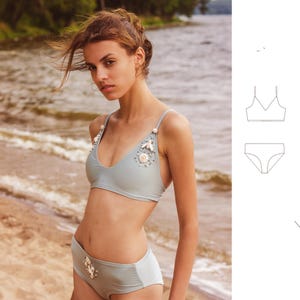 Pode incluir: Um biquíni azul claro com detalhes de conchas brancas. O top tem um decote em V e alças finas. A parte de baixo é de cintura alta e tem uma cintura larga.