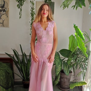 May include: A woman wearing a pink, sheer, sleeveless dress with lace detailing. The dress has a fitted bodice and a flowing skirt. The woman is standing in a room with plants.