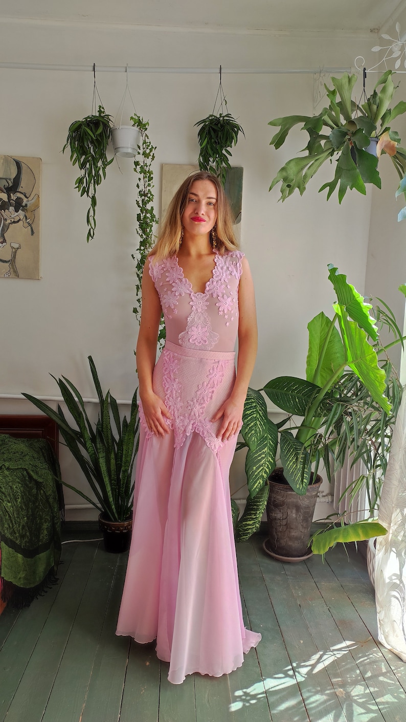 May include: A woman wearing a pink, sheer, sleeveless dress with lace detailing. The dress has a fitted bodice and a flowing skirt. The woman is standing in a room with plants.
