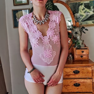 Könnte beinhalten: Ein durchsichtiger rosa Body mit floralen Spitzendetails. Der Body hat einen tiefen V-Ausschnitt und wird mit weißen Shorts kombiniert. Eine Statement-Halskette mit großen, hellfarbigen Steinen wird getragen. Das Bild ist in Innenräumen aufgenommen.