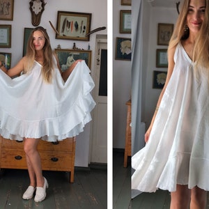 May include: A white, sleeveless dress with a V-neck and a tiered, ruffled hem. The dress is made of a lightweight fabric and falls to mid-thigh. The model is wearing white loafers.