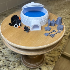 May include: A white plastic igloo with a blue interior and a small opening. Two black and white penguin figurines are next to the igloo. There are also several gray plastic castle and sandcastle toys.