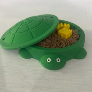 May include: A green turtle-shaped sandbox with a lid. The sandbox is filled with brown sand and a yellow plastic castle toy.