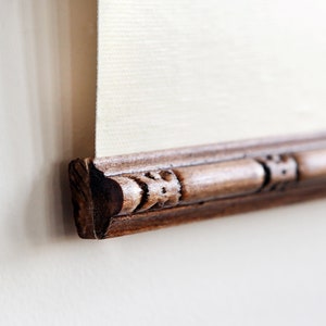 May include: Close-up of the bottom edge of a framed artwork. The frame is made of dark wood with an ornate design.