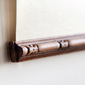 May include: Close-up of the bottom edge of a wooden frame with intricate carvings. The frame is dark brown and has a light brown finish.