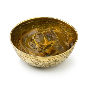 K&ouml;nnte beinhalten: Eine runde, goldfarbene Metallschale mit einem gepr&auml;gten Muster aus stilisierten Figuren und Blumenmotiven an der Au&szlig;enseite. Die Schale ist mit einer dicken, viskosen, dunkel bernsteinfarbenen Substanz gef&uuml;llt, die eine gl&auml;nzende Oberfl&auml;che hat.
