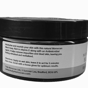 K&ouml;nnte beinhalten: Ein schwarzer zylindrischer Tiegel mit marokkanischer schwarzer Seife und einem wei&szlig;en Etikett. Der Text auf dem Etikett lautet: Moisturise and nourish your skin with this natural Moroccan Black Soap. Rich in vitamin E along with an Antimicrobial agent, it helps remove impurities and dead skin, leaving you feeling refreshed and revitalised. Directions: Apply on wet skin, leave it to act for 5 minutes then rinse. Scrub with a Kessa glove for optimum results.