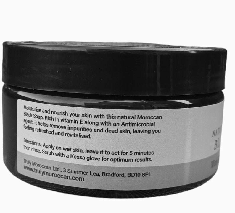K&ouml;nnte beinhalten: Ein schwarzer zylindrischer Tiegel mit marokkanischer schwarzer Seife und einem wei&szlig;en Etikett. Der Text auf dem Etikett lautet: Moisturise and nourish your skin with this natural Moroccan Black Soap. Rich in vitamin E along with an Antimicrobial agent, it helps remove impurities and dead skin, leaving you feeling refreshed and revitalised. Directions: Apply on wet skin, leave it to act for 5 minutes then rinse. Scrub with a Kessa glove for optimum results.
