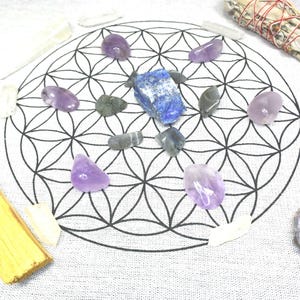 May include: A collection of crystals and spiritual items arranged on a white cloth with a black geometric design. The arrangement includes amethyst crystals, a blue lapis lazuli stone, sage, and palo santo.
