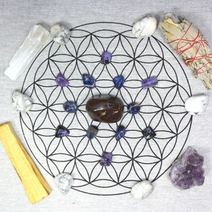 May include: A collection of crystals and spiritual items arranged on a white cloth with a black geometric design. The arrangement includes amethyst, selenite, sage, and palo santo, ideal for meditation and energy work.