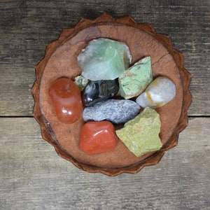 May include: A collection of colorful gemstones, including red, green, and gray varieties, are arranged in a decorative, brown, scalloped-edge bowl. The bowl sits on a rustic, weathered wooden surface, creating a natural and earthy aesthetic.