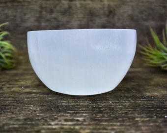 May include: A white selenite bowl, a type of crystal known for its calming energy, sits on a wooden surface. The bowl is smooth and polished, with a slightly rounded shape.