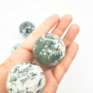 May include: A hand holds several round, polished gemstones. The stones are primarily white with green and black inclusions, creating a speckled pattern. The gemstones are spherical, and the background is plain white.