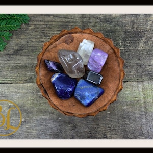 May include: A collection of polished gemstones in a small, brown, leather-like bowl. The stones vary in color, including deep blues, purples, and clear crystals. The bowl sits on a weathered wooden surface, with green leaves in the background.