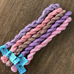 May include: Five skeins of embroidery floss in shades of pink, purple, and brown. The floss is wrapped around itself and the ends are tied together. The skeins are labeled "Gumnut Yarns "Buds".