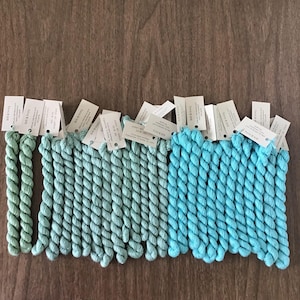 May include: An assortment of colorful embroidery floss in shades of green and blue. Each bundle is labeled with a small white tag. The floss is arranged on a wooden surface, showcasing the different hues and textures of the thread.