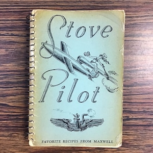 May include: A vintage cookbook titled "Stove Pilot: Favorite Recipes from Maxwell" with a blue and white cover featuring a cartoon illustration of a woman riding a jet plane.