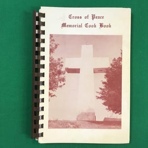 May include: A vintage cookbook with a brown spine and a white cover. The cover features a photo of a large white cross against a red sky. The title of the book is "Cross of Peace Memorial Cook Book".