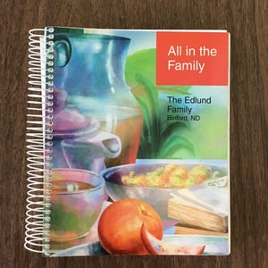 May include: A spiral-bound book titled All in the Family, The Edlund Family, Binford, ND. The cover features a colorful illustration of a pitcher, a bowl of food, an orange, and stacked books, with a prominent red rectangle in the upper right corner.