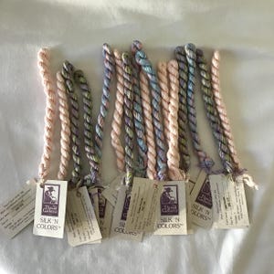 May include: A collection of silk threads in various pastel colors, including pink, blue, and green. Each thread is twisted and has a tag attached with the "Silk 'N Colors" logo and product information. The threads are arranged on a white surface.