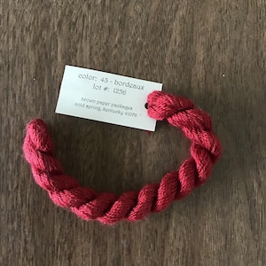May include: A skein of red embroidery floss, color 43-bordeaux, is coiled on a wooden surface. A small white tag attached to the floss displays the color name, lot number, and company information.