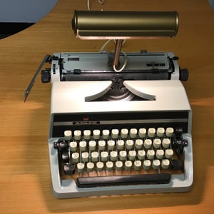 May include: A white vintage Adler typewriter with a black keyboard and a gold-colored lamp attached to the top. The typewriter has a black and white label that says "ADLER" on the front.