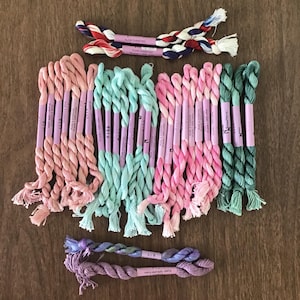 May include: A collection of colorful embroidery floss in various shades of pink, green, and blue, arranged on a wooden surface. The floss is neatly bundled with labels, showcasing a variety of hues for crafting and embroidery projects.