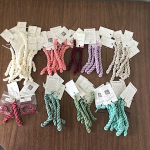 May include: A collection of colorful embroidery floss bundles in various shades, including white, pink, burgundy, purple, beige, teal, and green. Each bundle is accompanied by a tag. The floss is arranged on a brown surface.