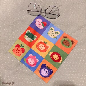 Puede incluir: Un pa&ntilde;o de limpieza de microfibra colorido con un patr&oacute;n de caras de animales con tem&aacute;tica de frutas. El pa&ntilde;o es cuadrado y presenta una variedad de colores, incluyendo rosa, verde, naranja y azul. Un par de gafas descansa sobre el pa&ntilde;o.