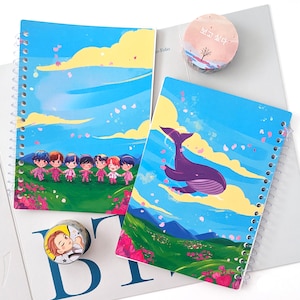 May include: Two spiral bound notebooks with colourful illustrations. One notebook features a blue sky with pink flowers and a group of cartoon characters. The other notebook features a blue whale swimming in a blue sky with pink flowers. The text "보고 싶다" is on a pink and white washi tape.