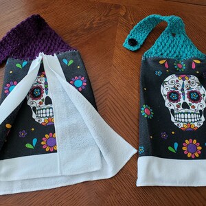 Pu&ograve; includere: Due asciugamani da cucina con sfondo nero e un design colorato di teschio di zucchero. Gli asciugamani hanno un bordo bianco e un retro in spugna bianca. Un asciugamano ha un'ansa di sospensione in crochet viola e l'altro ha un'ansa di sospensione in crochet turchese.