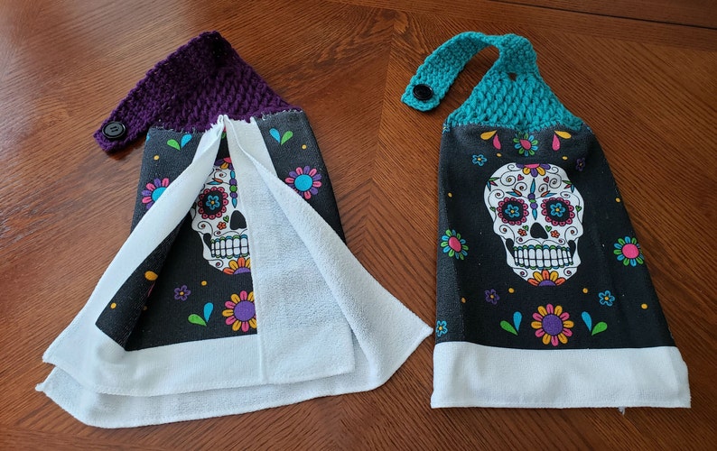 Pu&ograve; includere: Due asciugamani da cucina con sfondo nero e un design colorato di teschio di zucchero. Gli asciugamani hanno un bordo bianco e un retro in spugna bianca. Un asciugamano ha un'ansa di sospensione in crochet viola e l'altro ha un'ansa di sospensione in crochet turchese.