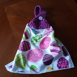 May include: Purple crocheted hanging loop with a button on a white kitchen towel with a colorful Easter egg pattern.