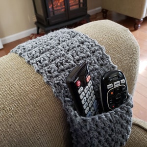 May include: Gray crocheted armrest cover with two black remote controls tucked inside. The armrest cover is on a beige couch.