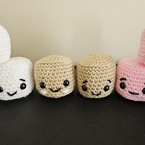 May include: A collection of handmade crochet plushies in white, tan, and pink. Each plushie has a smiling face with black eyes and a stitched mouth. The white plushies have pink cheeks. The plushies are arranged on a dark surface.