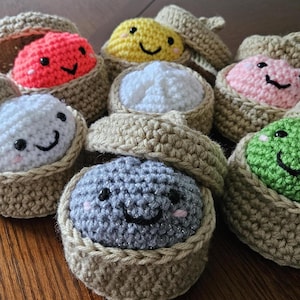 May include: A collection of seven small, round, crocheted plush toys, each nestled inside a tan, acorn-shaped container with a removable lid. The plush toys feature various colors including red, yellow, white, pink, green, and gray, all with embroidered faces.