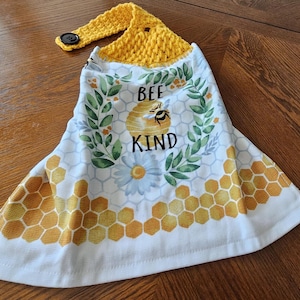 May include: A hanging kitchen towel with a yellow crocheted top and a button closure. The white towel features a bee and honeycomb design with the words "BEE KIND". The towel has a honeycomb pattern along the bottom edge.