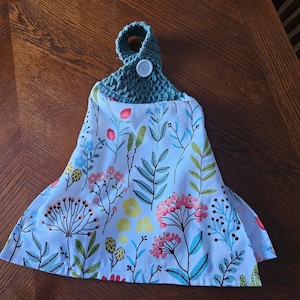 May include: A teal crocheted dish towel holder with a white button. The towel is white with a floral pattern in shades of pink, green, and yellow.
