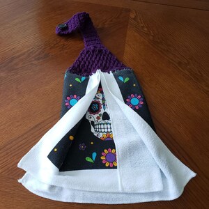 Pu&ograve; includere: Anello di sospensione in crochet viola con un bottone su un asciugamano da cucina nero e bianco con un design di teschio di zucchero colorato.