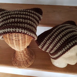 May include: Two brown and beige striped crocheted hats on display stands. The hats are made with a textured stitch and have a simple design.