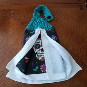 Pu&ograve; includere: Un anello di sospensione in crochet turchese con un bottone nero &egrave; attaccato a un asciugamano da cucina bianco su sfondo nero. L'asciugamano presenta un design colorato di teschio di zucchero.