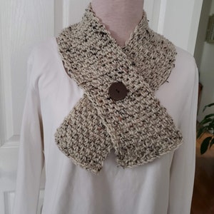 May include: A hand-crocheted scarf in a neutral color palette, featuring flecks of brown and black. The scarf is secured with a large, round, brown button. The textured stitch and button detail create a cozy, handmade aesthetic.