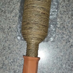 May include: A decorative bottle-shaped item with a terracotta-colored pointed base and a cylindrical top wrapped in natural-colored twine. The item appears to be a plant watering tool.