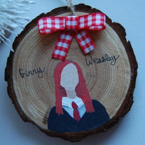 May include: A wooden slice ornament featuring a hand-painted illustration of a character with red hair, a white shirt, and a red tie. The name "Ginny Weasley" is written above the illustration. A red and white checkered bow adorns the top.