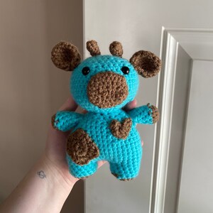 May include: A crocheted blue and brown cow plush toy. The cow has brown ears, a brown nose, and brown spots on its body.