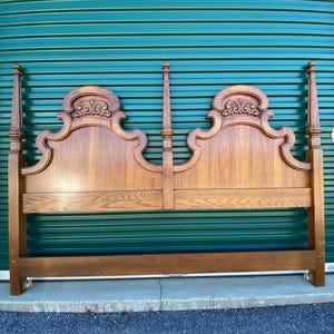 May include: A wooden headboard with a classic design. The headboard is made of light brown wood and features ornate carvings on the top portion. It has four tall posts and a low base, suitable for a bed frame.