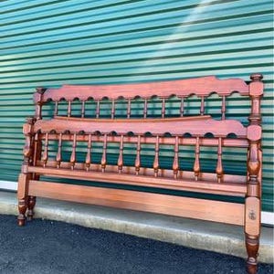 Puede incluir: Un marco de cama de madera con cabecero y piecero. El marco está hecho de madera de tonos cálidos y presenta husillos decorativos y detalles tallados. El marco de la cama está contra una pared de color verde azulado.