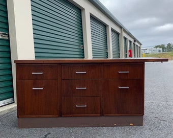 Può includere: Un mobile in legno marrone scuro con sei cassetti e maniglie argentate. Il mobile ha una base marrone scuro e un piano piatto. Il mobile è di fronte a una fila di porte verdi di unità di stoccaggio.